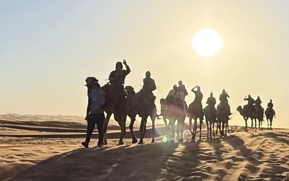 Riders on camels in silhouette against a low sun over the dunes