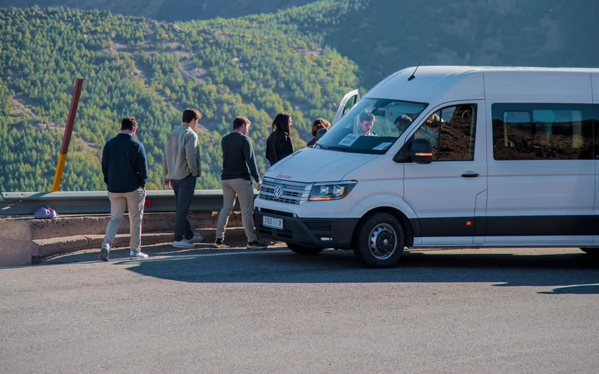 A minibus stopped at a roadside viewpoint with passengers looking out over a wooded slope